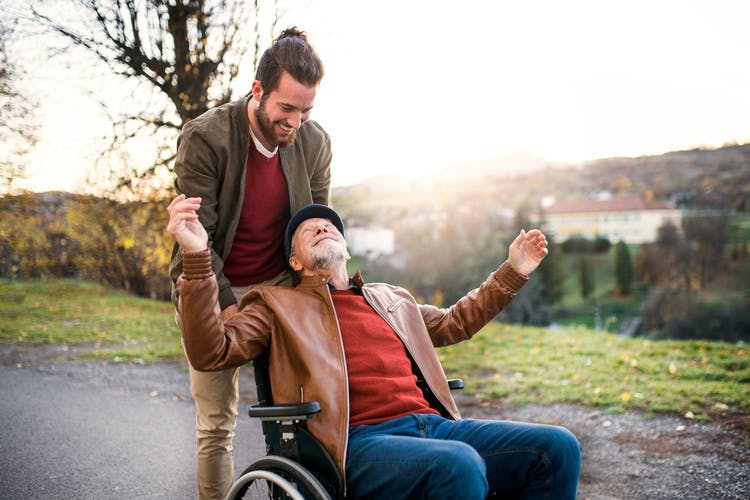 son pushing wheelchair of elderly father who has hemiplegia and hemiparesis through the park