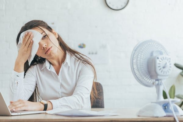 woman wiping head with cloth sitting in front of electric fan in office because she struggles with excessive sweating after brain injury