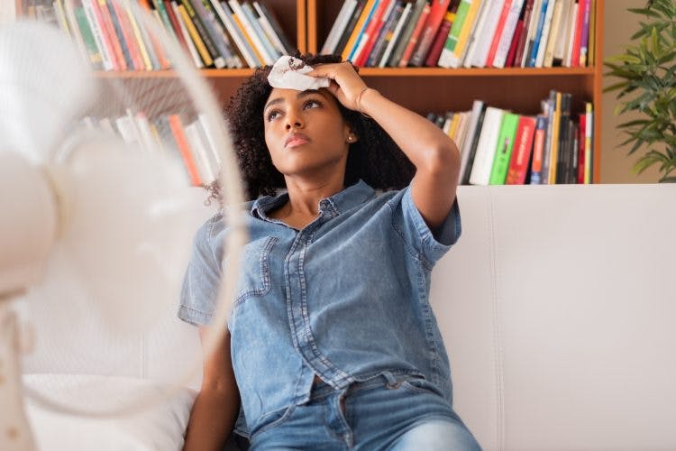woman sitting on couch in front of a fan, holding a cold cloth on her forehead, dealing with TBI and heat sensitivity