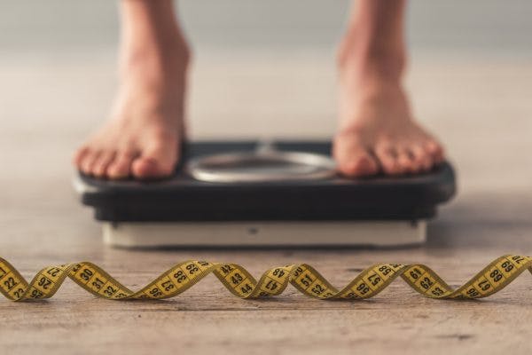 Cropped image of woman feet standing on weigh scales to measure her weight gain after brain injury. A tape measure lies on the floor in the foreground