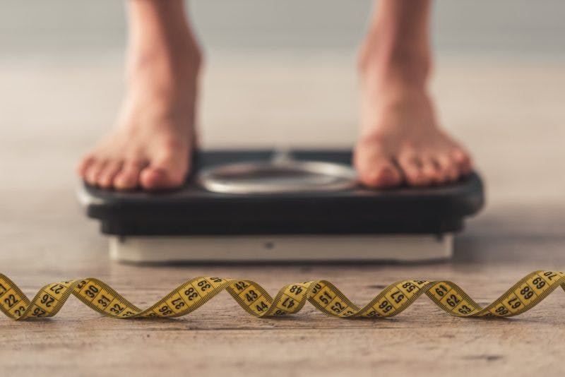 Cropped image of woman feet standing on weigh scales to measure her weight gain after brain injury. A tape measure lies on the floor in the foreground